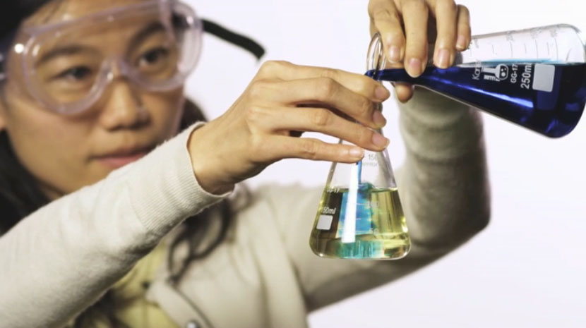 A woman wearing goggles poring blue liquid from a beaker into a test tube
